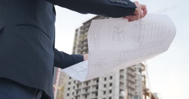 Um jovem arquiteto construtor inspeciona a construção do edifício. Um engenheiro olha para os desenhos contra o fundo da construção do edifício na rua contra o — Vídeo de Stock