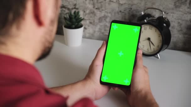 En man vid ett vitt skrivbord sitter med en mobiltelefon i sina händer med en grön skärm. sveper och kranar på smartphones chromakey screen i närbild. Färdig video för insättning och nyckelring — Stockvideo