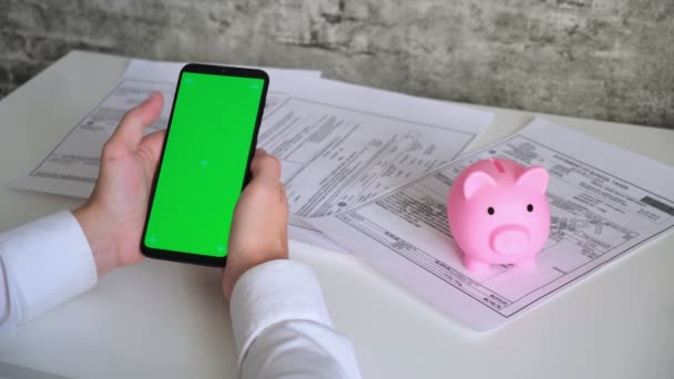 Telefone com tela verde e recibos de pagamento piggy Bank, documentos fiscais no topo do fundo branco View. smartphone chromakey rouba em direções diferentes. espaço de cópia — Vídeo de Stock