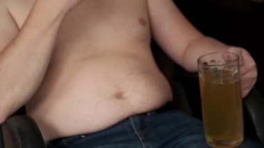 Şişman adam cips yiyor ve bira içiyor. El bira göbeğini kaşır. Zararlı yiyeceklerin bira göbeği. Kötü yemek, oburluk. Yakın plan..