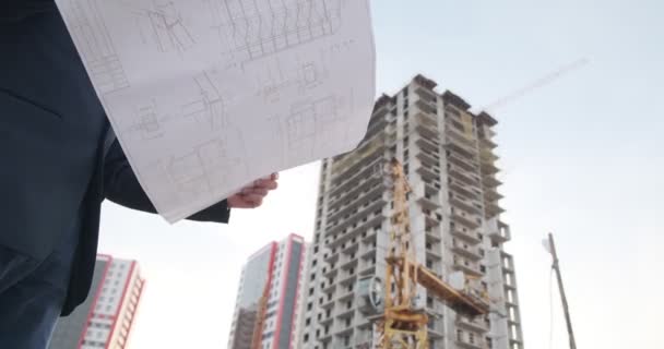 Um jovem arquiteto construtor inspeciona a construção do edifício. Um engenheiro olha para os desenhos contra o fundo da construção do edifício na rua contra o — Vídeo de Stock
