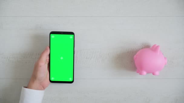 Teléfono con una pantalla cromakey verde sobre un fondo blanco con una vista superior alcancía rosa. espacio copia smartphone pasa en diferentes direcciones — Vídeos de Stock