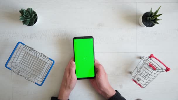 Телефон с зеленым цветным экраном и корзиной для покупок на белом фоне. Вид смартфона в руках крупного плана — стоковое видео