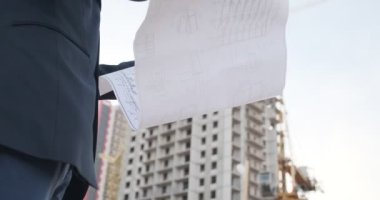 Genç bir erkek mimar binanın inşaatını denetliyor. Bir mühendis, binanın arka planındaki çizimlere bakıyor.