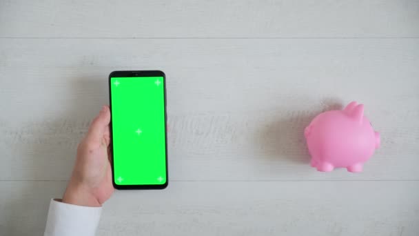 Teléfono con una pantalla cromakey verde sobre un fondo blanco con una vista superior alcancía rosa. espacio copia smartphone pasa en diferentes direcciones — Vídeos de Stock