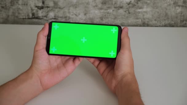 En man vid ett vitt skrivbord sitter med en mobiltelefon i sina händer med en grön skärm. sveper och kranar på smartphones chromakey screen i närbild. Färdig video för insättning och nyckelring — Stockvideo