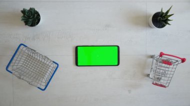 Yeşil renkli ekranlı bir telefon ve beyaz arka planda alışveriş arabası. Akıllı telefonun tepesinden görüntüle