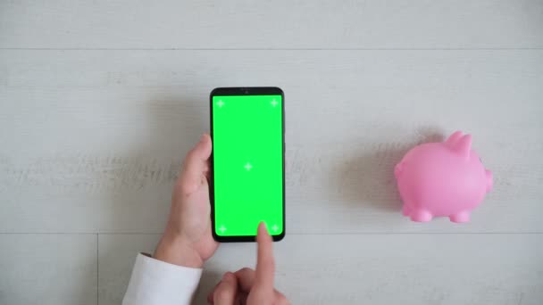 Teléfono con una pantalla cromakey verde sobre un fondo blanco con una vista superior alcancía rosa. espacio copia smartphone pasa en diferentes direcciones — Vídeos de Stock