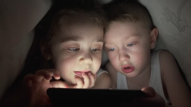 Un niño y una niña felices se acuestan en la cama debajo de una manta y juegan en un teléfono en un juego en la oscuridad. hermano y hermana cara del niño está iluminado por un niño monitor brillante bajo las cubiertas niño adicto a la computadora — Vídeos de Stock