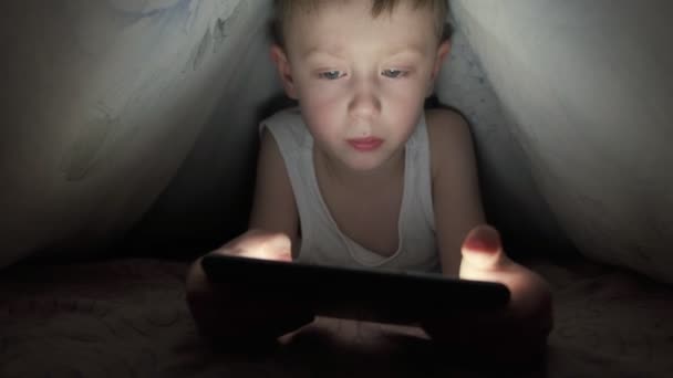 Um menino feliz está deitado na cama sob um cobertor e joga no celular em um jogo no escuro. O rosto da criança é iluminado por um monitor brilhante. Imagens 4k de alta qualidade. — Vídeo de Stock