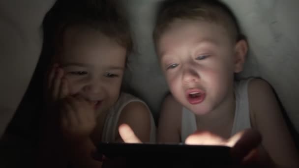 Un niño y una niña felices se acuestan en la cama debajo de una manta y juegan en un teléfono en un juego en la oscuridad. hermano y hermana cara del niño está iluminado por un niño monitor brillante bajo las cubiertas niño adicto a la computadora — Vídeos de Stock