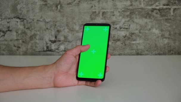 Mężczyzna przy białym biurku siedzi z telefonem komórkowym w rękach z zielonym ekranem. przesuwa i stuka na ekranie Chromakey smartfonów w zbliżeniu. gotowe wideo do wkładania i kluczenia — Wideo stockowe