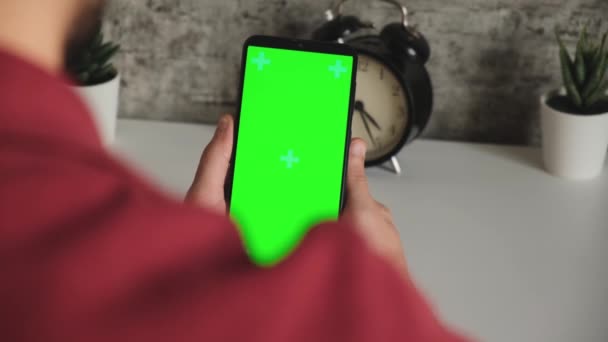 Un uomo a una scrivania bianca è seduto con un telefono cellulare in mano con uno schermo verde. scorre e tocca lo schermo del cromakey degli smartphone in primo piano. video pronto per l'inserimento e la digitazione Video Stock