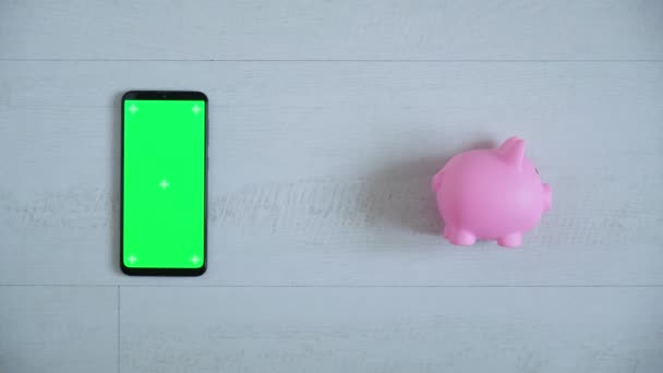 ピンクのピギーバンクのトップビューを持つ白い背景に緑のクロマキー画面を持つ電話。スマートフォンのコピースペースは — ストック動画