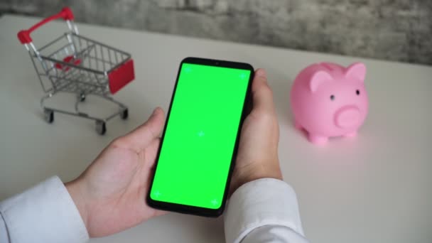Telefon mit einem Chromakey-Bildschirm in den Händen einer Nahaufnahme. von oben Smartphone Sparschwein und Warenkorb Kopierraum auf weißem Hintergrund — Stockvideo