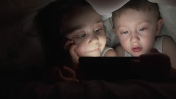 Um menino e uma menina felizes jazem na cama sob um cobertor e jogam em um telefone em um jogo no escuro. irmão e irmã rosto da criança é iluminado por um menino monitor brilhante sob as capas Criança viciada em computador — Vídeo de Stock