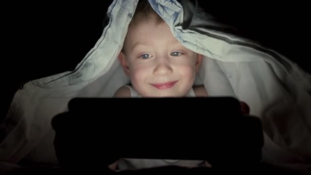 Un ragazzo felice si trova a letto sotto una coperta e gioca al cellulare in un gioco al buio. Il volto del bambino è illuminato da un monitor luminoso. Filmati 4k di alta qualità. Video Stock Royalty Free