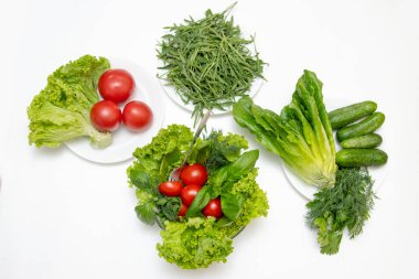 Top view of fresh vegetables tomatoes, cucumbers, parsley, dill, basil, rucola and green leaves for salad in plates on a white table.