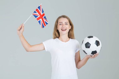 Büyük Britanya bayrağı ve elinde bir futbol topu olan genç bir kadın gri arka planda izole bir şekilde kameraya bakıyor. İngiliz kadın futbolu.