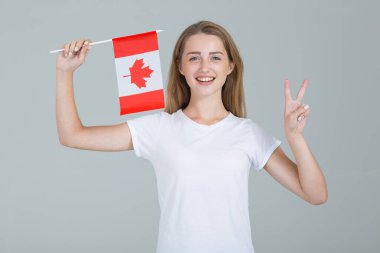 Kanada bayrağıyla güzel bir genç kadın ve iki parmağını bir şans işareti olarak gösteriyor. Gri arka planda izole edilmiş kameraya bakıyor..