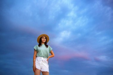 Gün batımında mavi bulutlu bir gökyüzünün arka planında hasır şapkalı güzel bir genç kadının portresi. Boşluğu kopyala.