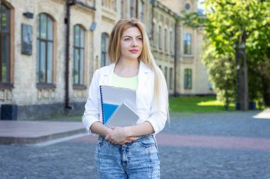 Ders kitaplarını tutan, üniversite arka planında duran genç öğrenci kadın portresi.