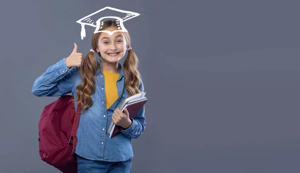 Schoolgirl girl with textbooks and graduate hat shows thumbs up. Studio photo on a gray background. Concept for text advertising about school and graduation.