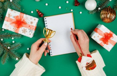 Beyaz kazaklı kadın elleri noel yeşili arka planda fotokopi aletiyle spiral not defterine yazıyorlar. Üstten bakan ilk kişi. Yeni yıl hediye listesi.