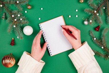 Yeni yıl kararları ya da hedeflerini spiral bir deftere yazan bir kadın. Noel süslemeleri ve fotokopi alanı olan yeşil bir arka plan. Noel Baba 'ya mektup ve alışveriş listesi.