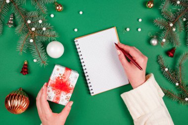 Yeni yıl kararları ya da hedeflerini spiral bir deftere yazan bir kadın. Noel süslemeleri ve fotokopi alanı olan yeşil bir arka plan. Noel Baba 'ya mektup ve alışveriş listesi.
