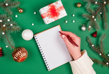 Yeni yıl kararları ya da hedeflerini spiral bir deftere yazan bir kadın. Noel süslemeleri ve fotokopi alanı olan yeşil bir arka plan. Noel Baba 'ya mektup ve alışveriş listesi.