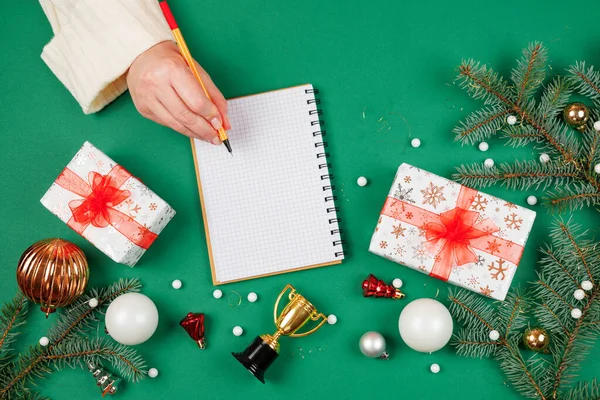 Beyaz kazaklı kadın elleri noel yeşili arka planda fotokopi aletiyle spiral not defterine yazıyorlar. Üstten bakan ilk kişi. Yeni yıl hediye listesi.