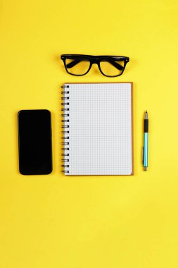 Sarı arka planda açık bir defter, kalem, gözlük ve akıllı telefonun üst görüntüsü. Dikey afiş. İş planlama, eğitim ve yaratıcı bloglar için çalışma alanı yatıyordu. Temiz model..