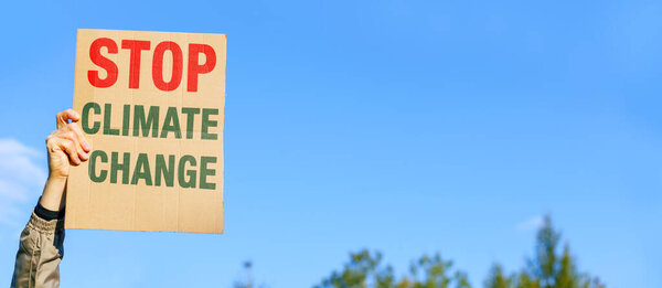 Male ecologist activist raises his hand and holds a sign reading STOP CLIMATE CHANGE against of nature and a blue sky background. Environmental protest against ecology issues.