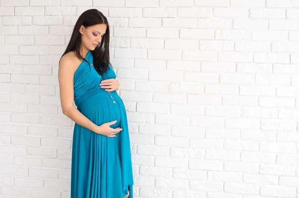 Красивая беременная женщина в бирюзовом платье на фоне белой кирпичной стены — стоковое фото