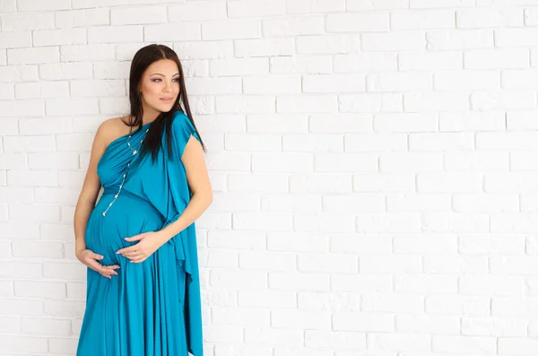 Beautiful pregnant woman in a turquoise dress on white brick wall background — Stock Photo, Image