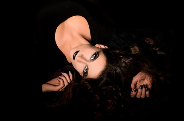 Lying young grey-eyed woman with professional art agressive makeup and wavy hair in decollete dress on black background