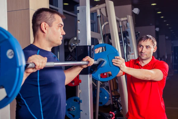 Deporte, fitness, trabajo en equipo, culturismo concepto de personas - hombre y entrenador personal con los músculos de flexión de la barra en el gimnasio —  Fotos de Stock