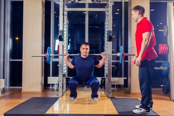 Deporte, fitness, trabajo en equipo, culturismo concepto de personas - hombre y entrenador personal con los músculos de flexión de la barra en el gimnasio —  Fotos de Stock