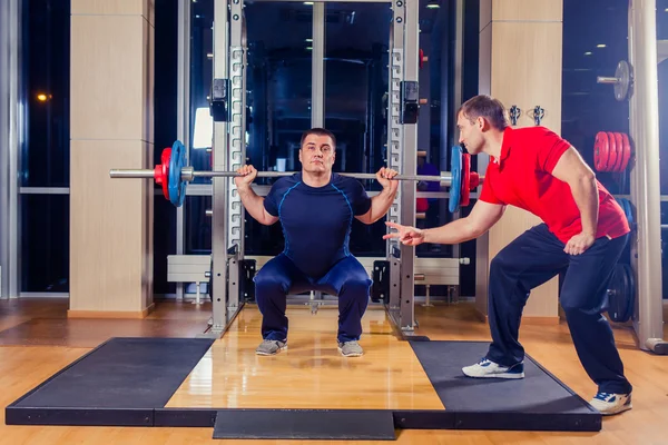 Deporte, fitness, trabajo en equipo, culturismo concepto de personas - hombre y entrenador personal con los músculos de flexión de la barra en el gimnasio —  Fotos de Stock