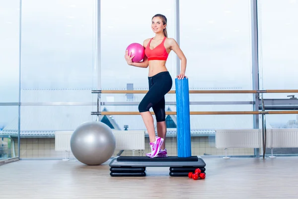 Chica de fitness, usando zapatillas de deporte, top rojo y pantalones negros, posando en el tablero de a pie con la pelota, el fondo del equipo deportivo, gimnasio — Foto de Stock