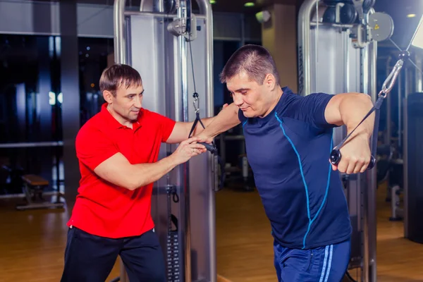Entrenador personal que ayuda a los hombres a ejercitarse en la sala de pesas en el gimnasio — Foto de Stock