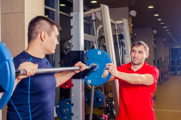 Sport, fitnesz, csapatmunka, testépítés emberek koncepció - ember és személyi edző a súlyzó hajlító izmok a tornaterem — Stock Fotó