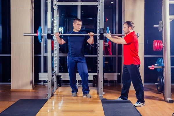 Esporte, fitness, trabalho em equipe, musculação conceito de pessoas homem e personal trainer com músculos de flexão barbell no ginásio — Fotografia de Stock