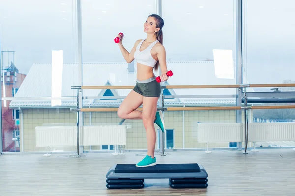 Happy young woman is doing exercises with dumbbells on step board.