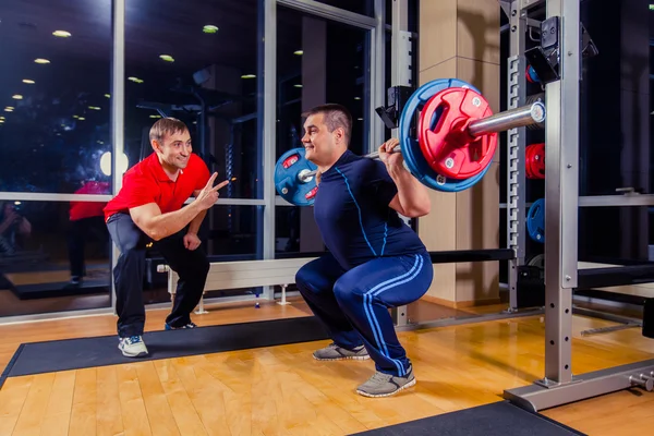 Deporte, fitness, trabajo en equipo, culturismo concepto de personas - hombre y entrenador personal con los músculos de flexión de la barra en el gimnasio —  Fotos de Stock