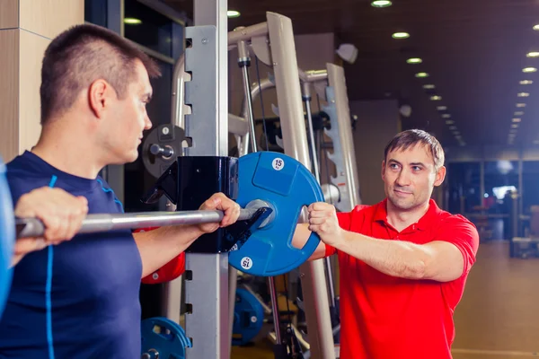 Deporte, fitness, trabajo en equipo, culturismo concepto de personas - hombre y entrenador personal con los músculos de flexión de la barra en el gimnasio —  Fotos de Stock