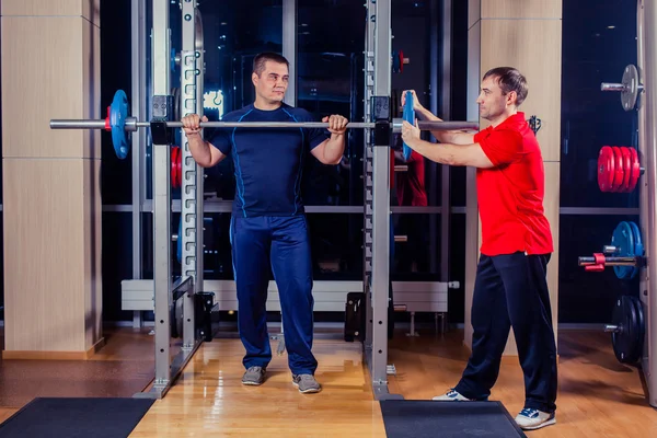 Deporte, fitness, trabajo en equipo, culturismo concepto de personas - hombre y entrenador personal con los músculos de flexión de la barra en el gimnasio — Foto de Stock