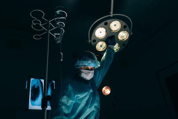 surgery, medicine and people concept - surgeon in mask adjusting lamp  operating room at hospital