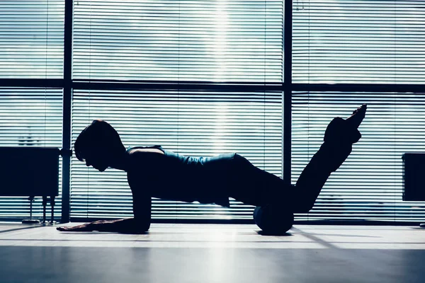 fitness, sport, training and lifestyle concept - woman doing pilates on the floor with foam roller contour of the body against the window